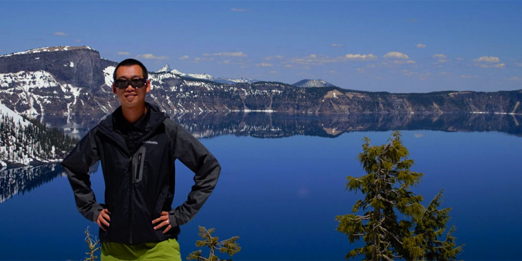 Man in sunglasses stands in front of lake surrounded by mountains.