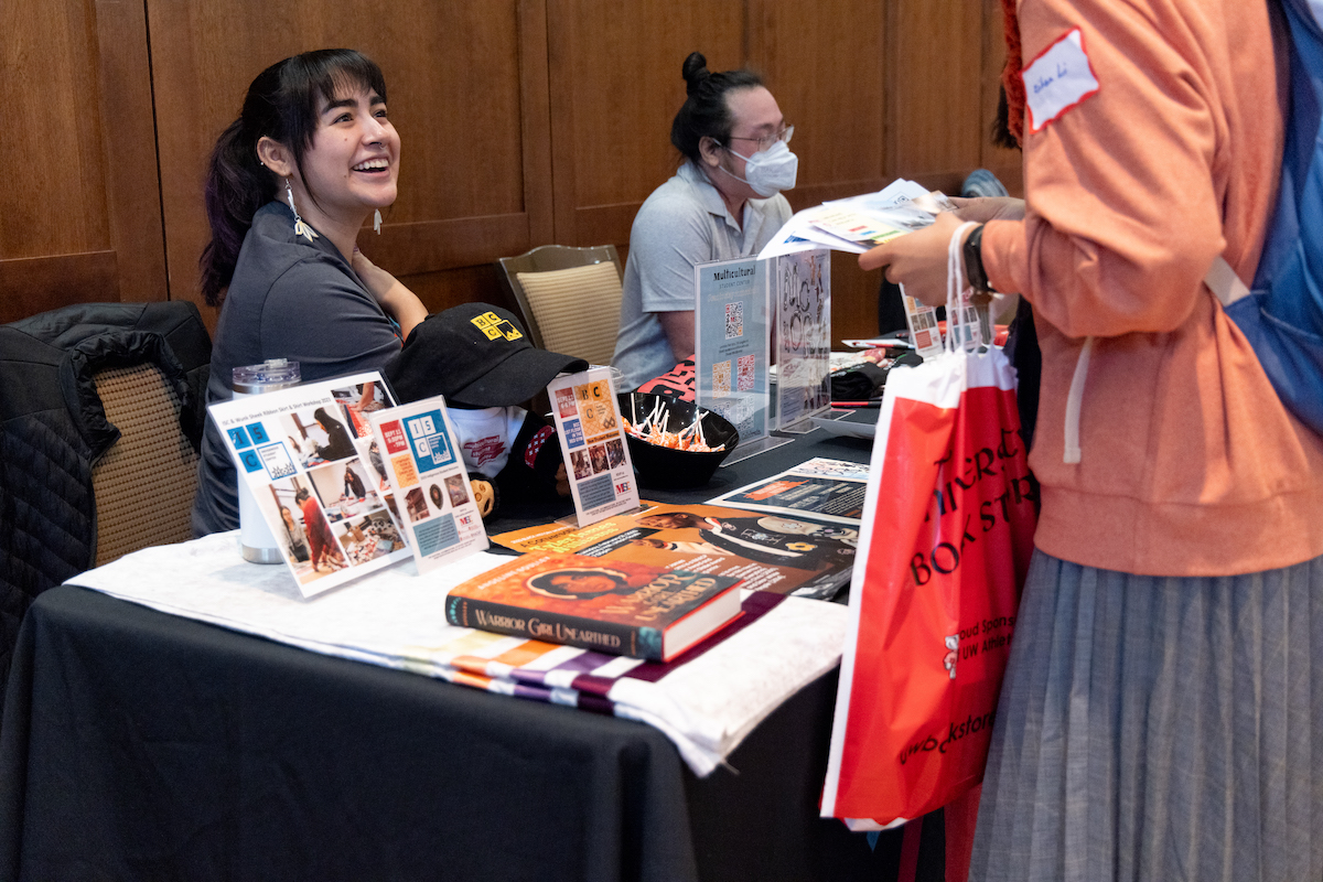 A person sits behind a booth covered in flyers, books, and giveaways and smiles while listening to someone else.