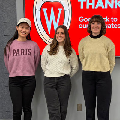 Three female Professional Masters students pose together in front of a screen with the UW–Madison crest.