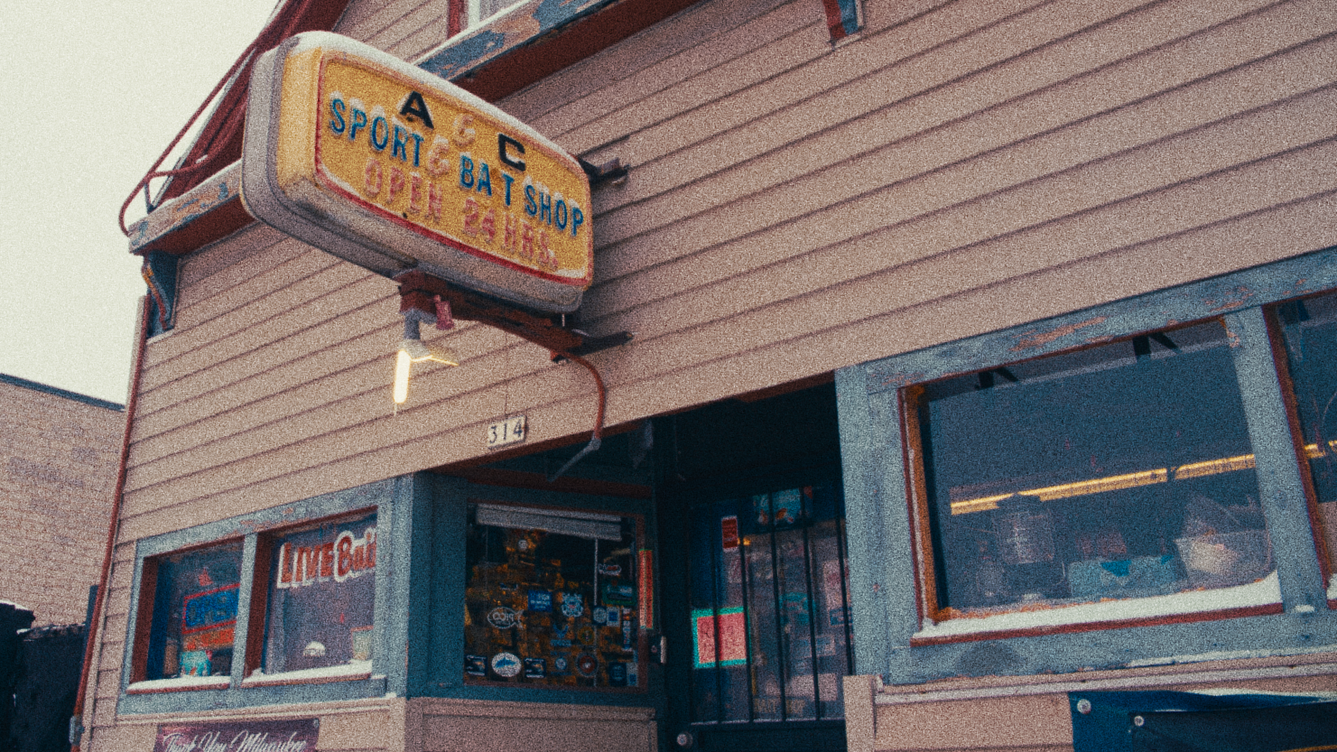 The outside of an old storefront has green wood around the windows filled with fishing gear and signs that read "open" and "live bait". A large plastic sign protrudes from the brown-sided building that reads "A & C Sport & Bait Shop Open 24 Hours"