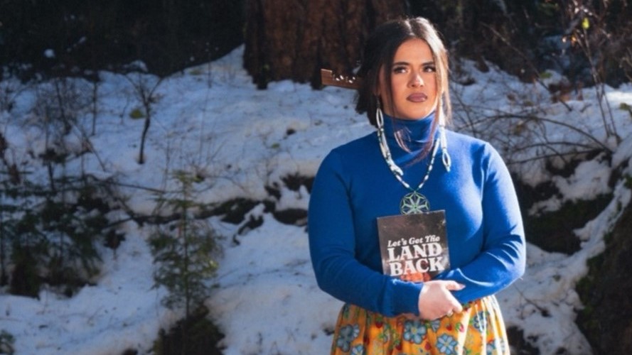 Tanikwah Lang stands outside and holds a book with the title "Let's Get The Land Back."