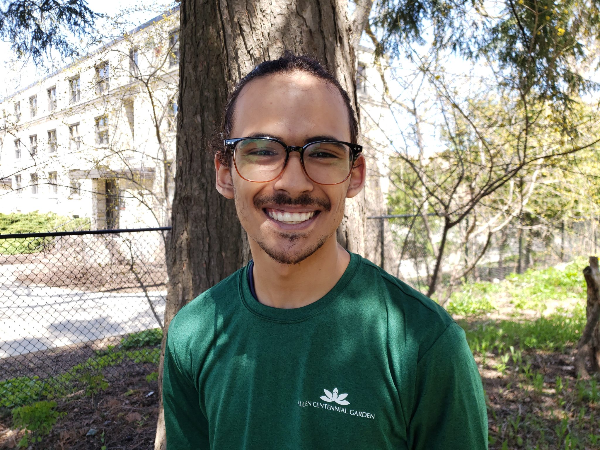Isaac Zaman smiles. He stands outside under a tree and wears a green Allen Centennial Garden t-shirt.