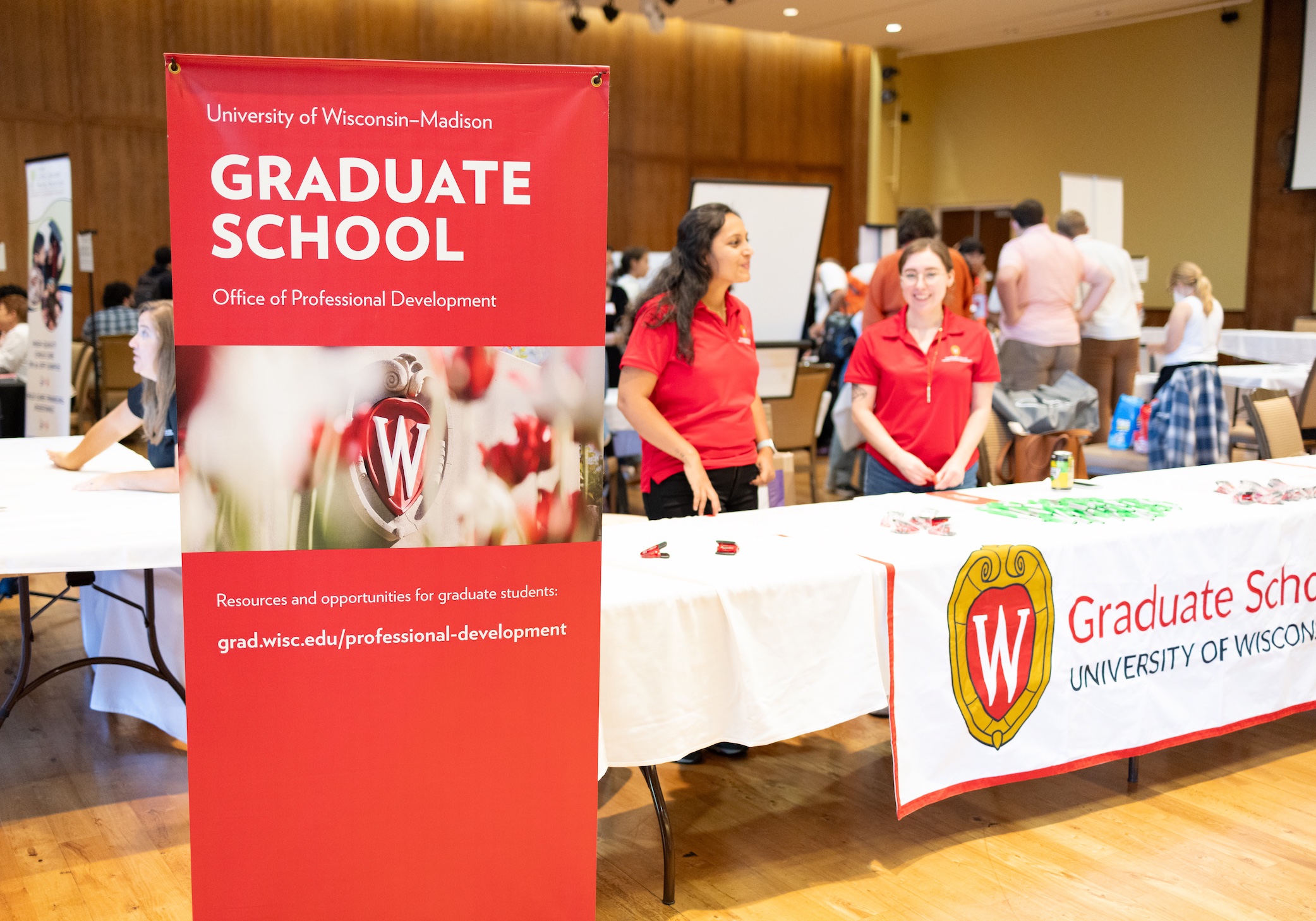 Two people in red polos stand at a table behind a banner that reads Graduate School.
