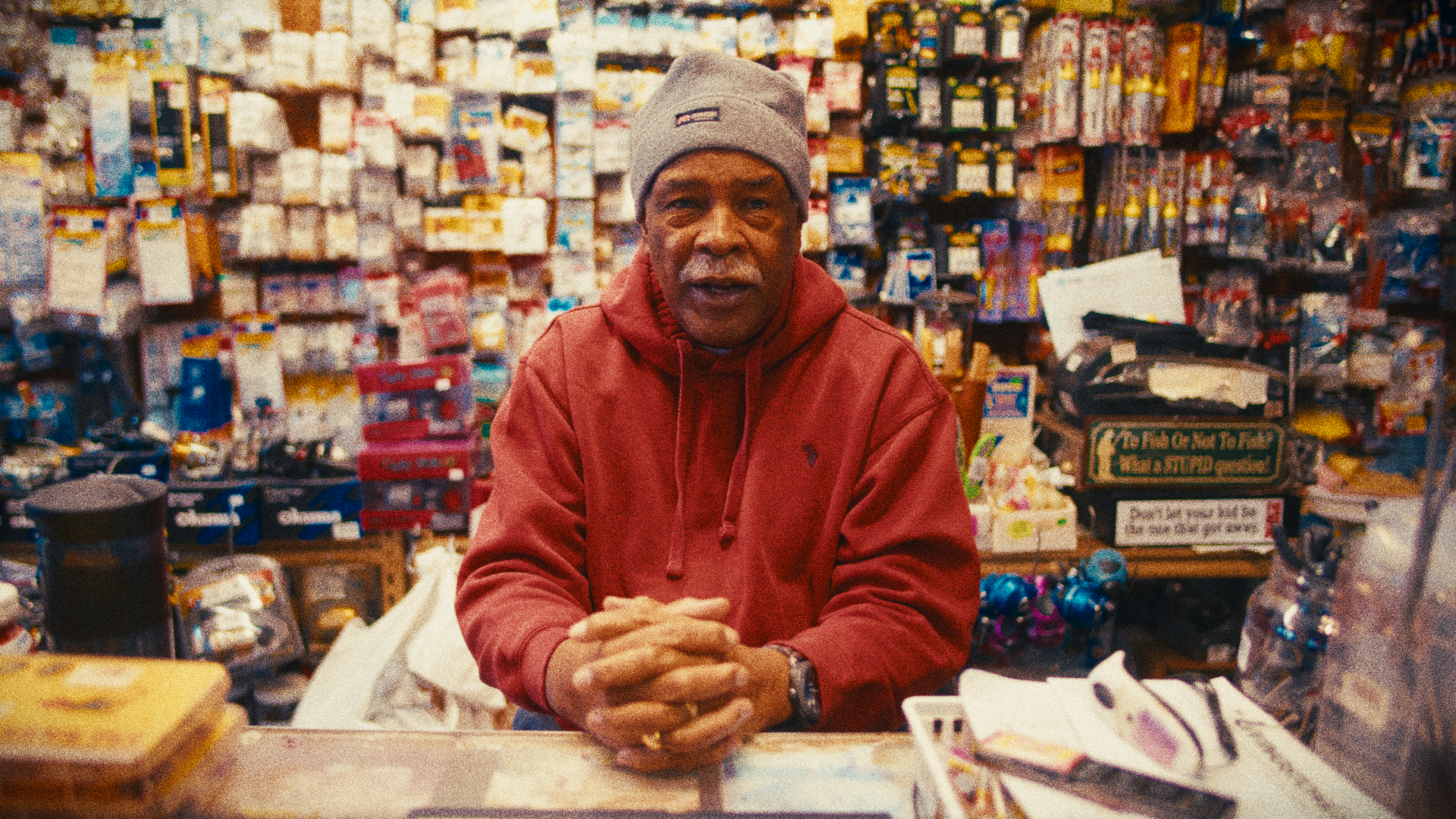 A man in a red hoodie wearing a gray stocking cap stands behind a counter with his fingers interlaced. The wall in the background is completely full of fishing lures and other fishing gear.