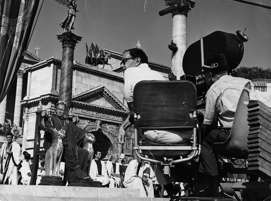 Director Joseph L. Mankiewicz seated next to a cameraman, with Rex Harrison and other actors on the Cleopatra set behind them.