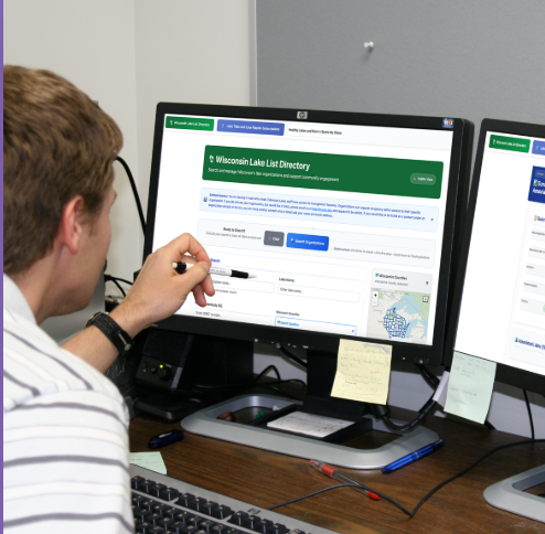 Man at a computer with two screens pointing to the left screen with a pen. On the screens are pages for the Wisconsin Lake List Directory.