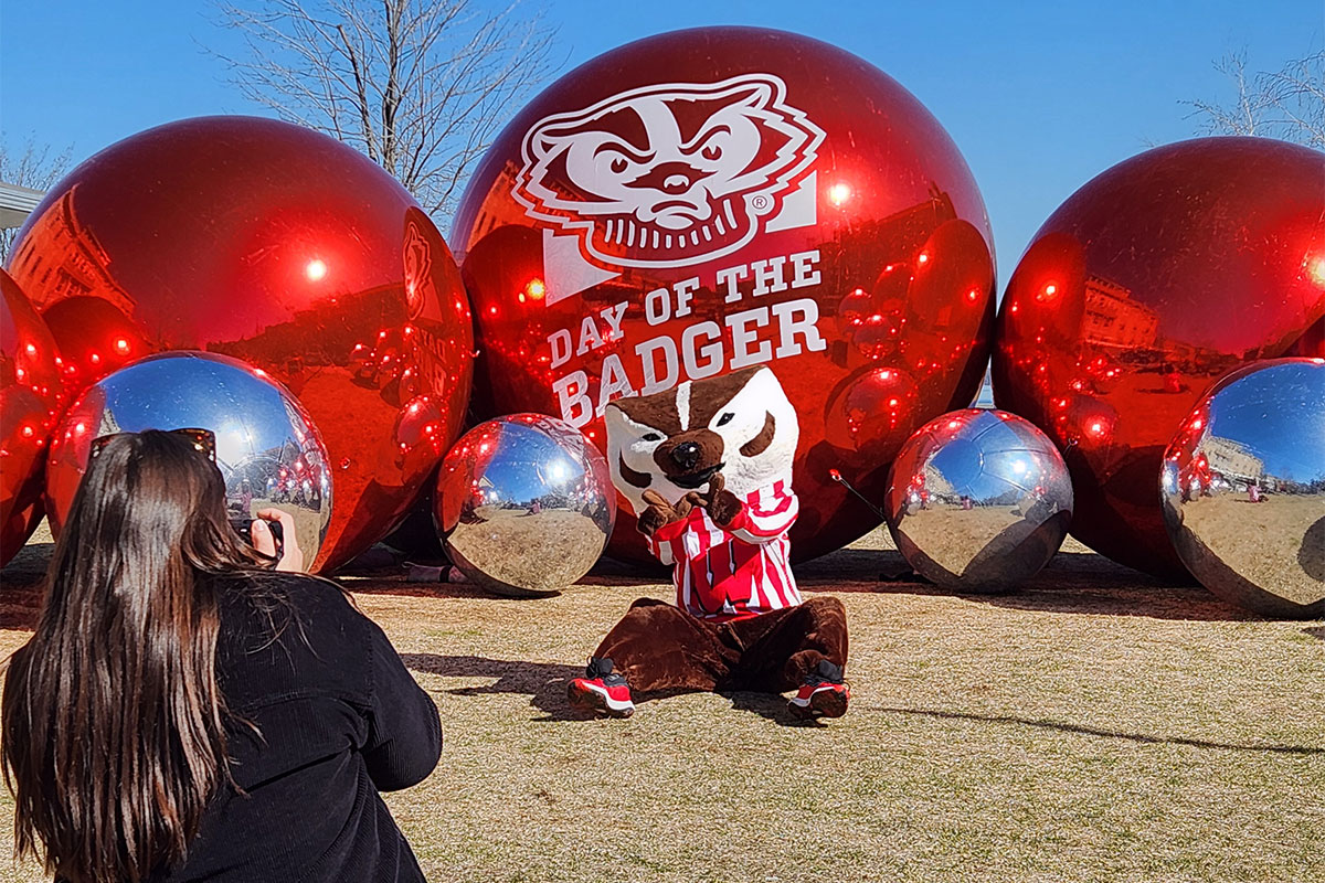 Bucky Badger getting their photo taken in front of the 2025 Day of the Badger Alumni Park display.