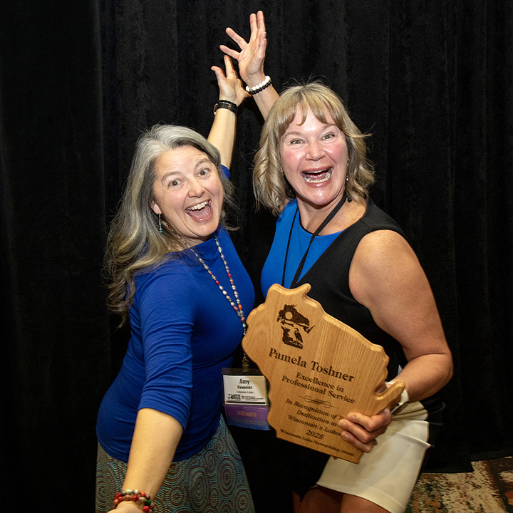Two smiling women celebrating an award shaped like the state of Wisconsin and cut out of wood.