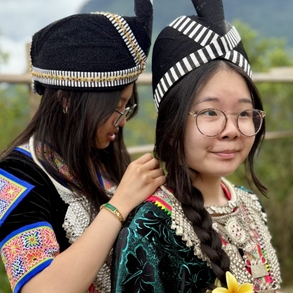 Two students stand in profile, both traditionally dressed in Hmong fashions, one standing behind the other pinning something on.