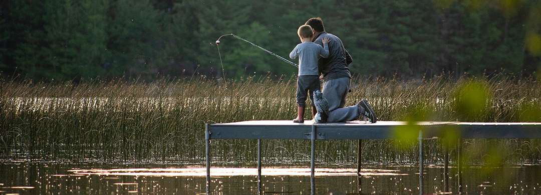 Young boy in red rain boots, long pants and long-sleeved shirt standing on a dock with his hand on his dad's shoulder, while dad is in jeans and a long-sleeved shirt kneeling on the dock with a fishing pole and a red and white bobber on the line. Pier is on a quiet lake with emergent vegetation and evergreens along the shore