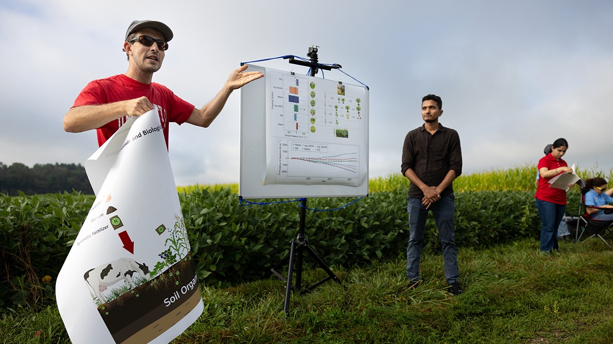 Steven Hall gestures to a poster in a field. People stand behind him and listen.
