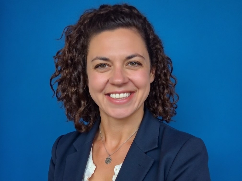Rachel Bouressa smiles. She wears a blue blazer and stands in front of a vibrant blue background.
