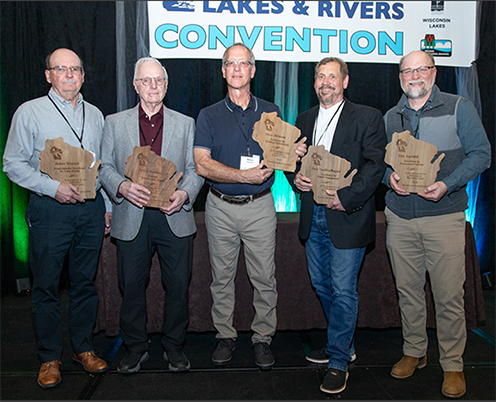 Five men dressed in business casual facing the camera and holding wooden awards in the share of the state of Wisconsin. In the background hangs a sign with the words "Lakes & Rivers Convention."