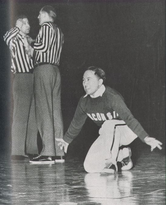 A black and white photo of a man in a UW cheerleading outfit kneeling on a gym floor