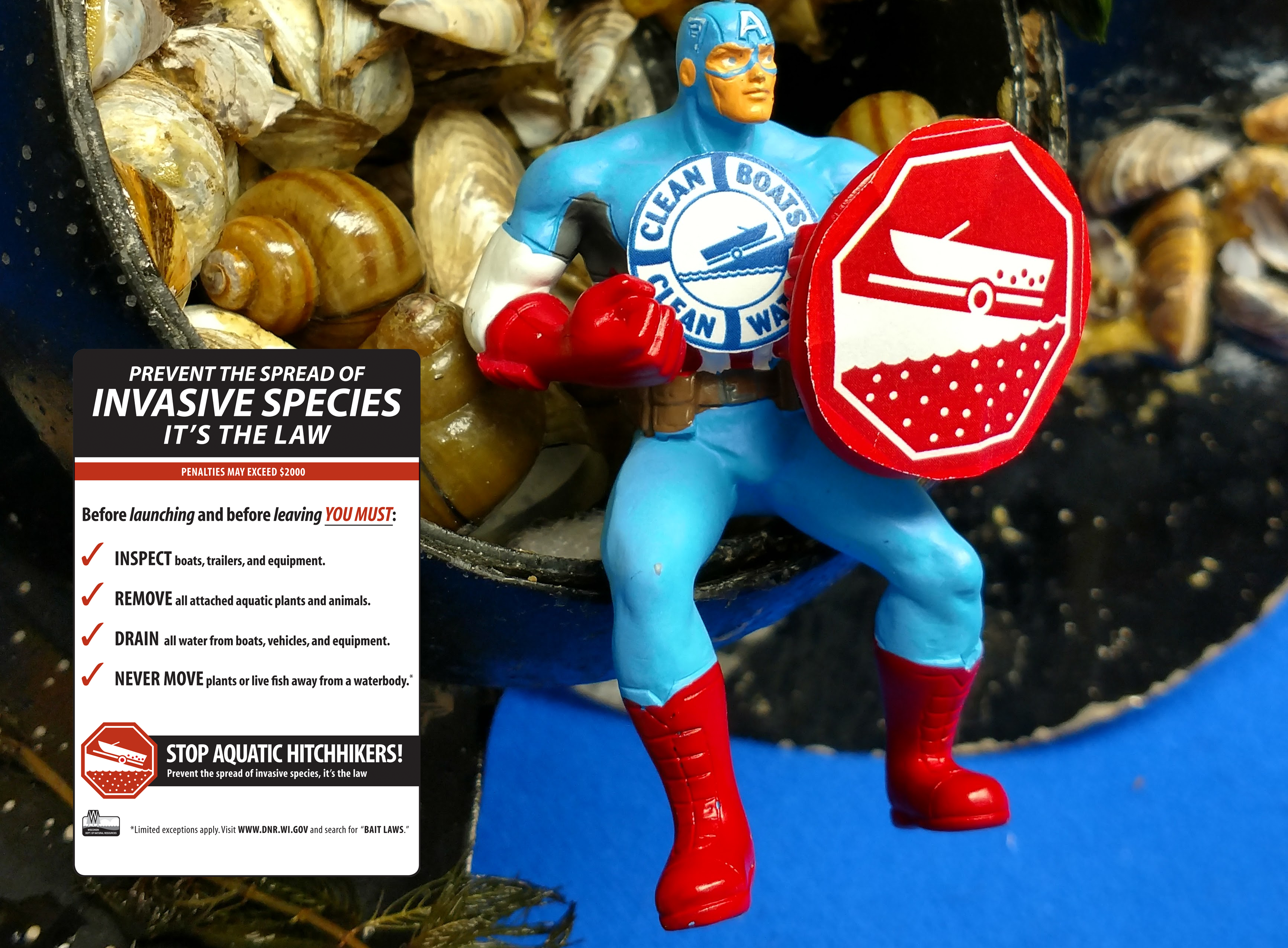 An action figure in a blue suit with red gloves and boots is holding a shield with the red "Stop Aquatic Hitchhikers!" logo while sitting in a boat propeller filled with aquatic invasive species - superimposed in the image is a sign reading "Prevent the spread of invasive species. It's the law"
