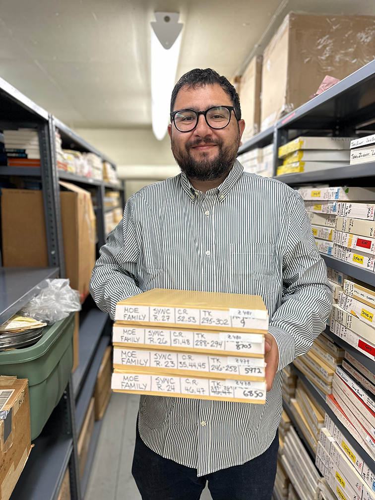 A smiling man holding four boxes of film stands in an aisle of shelves with film boxes and cans.