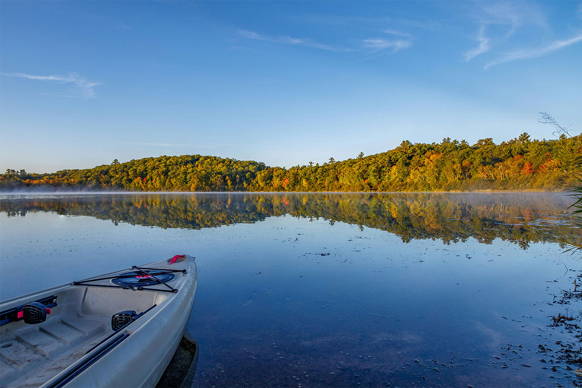 Front of a kayak looking out across a Wisconsin lake with autumn trees lining the opposite shore. 