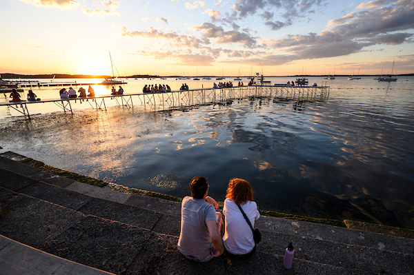 The sun sets over a glassy lake, casting light and shadows across a pier where people are seated.