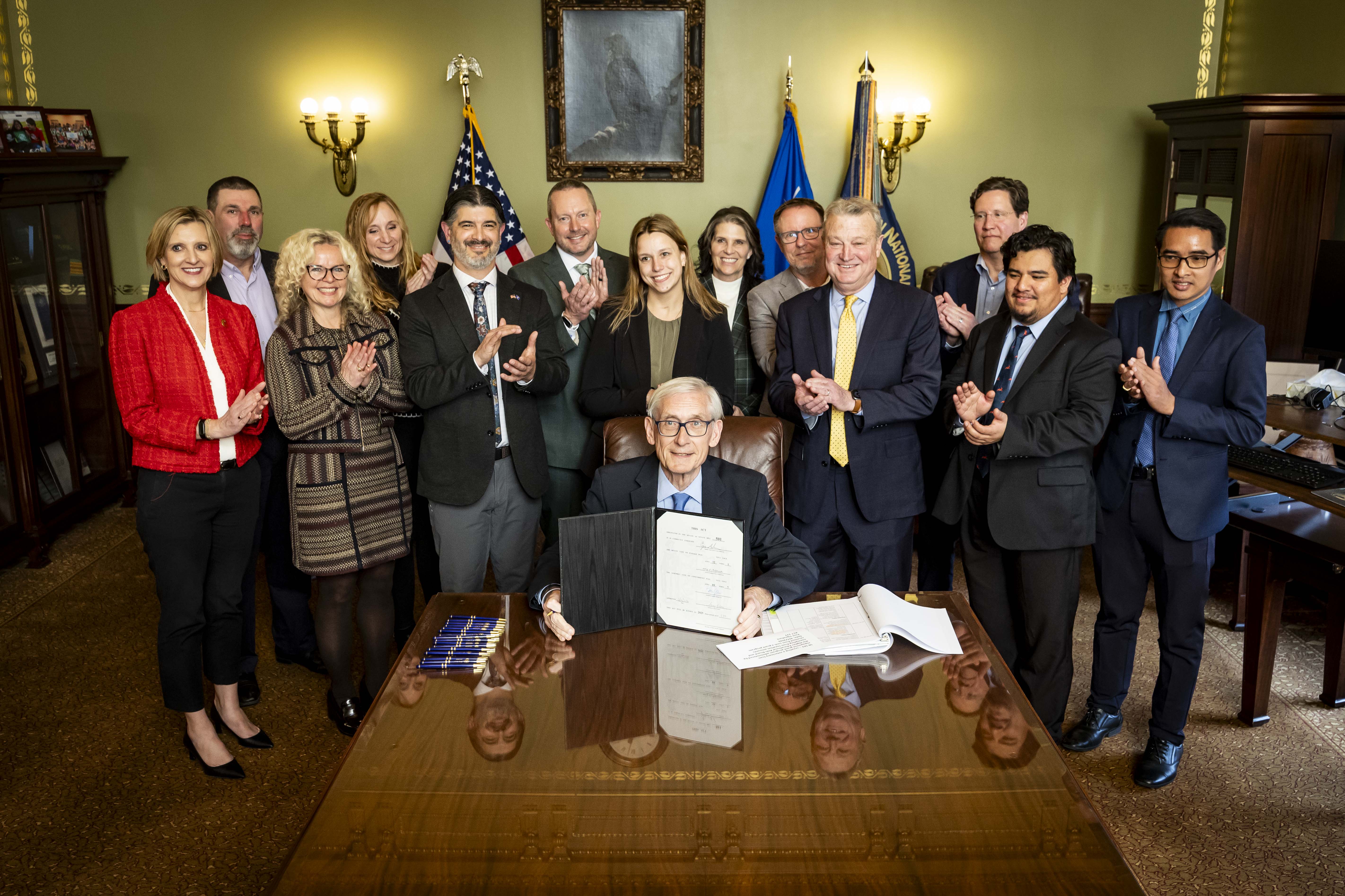 A group of people stands clapping behind a seated man holding a signed document, in a formal room with flags, conveying a celebratory mood.