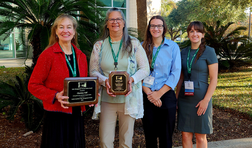 Four smiling women standing in front of palm trees holding award plaques