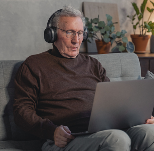 An older man with gray hair and glasses sits on a couch wearing headphones, using a laptop on his lap in a cozy living room surrounded by houseplants.