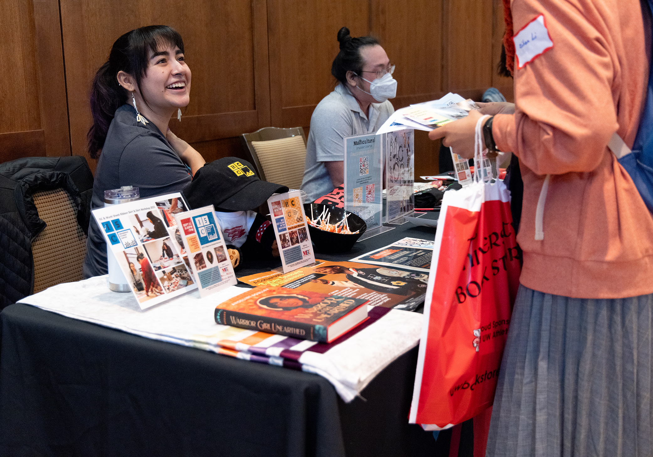 A person sits behind a booth covered in flyers, books, and giveaways and smiles while listening to someone else.