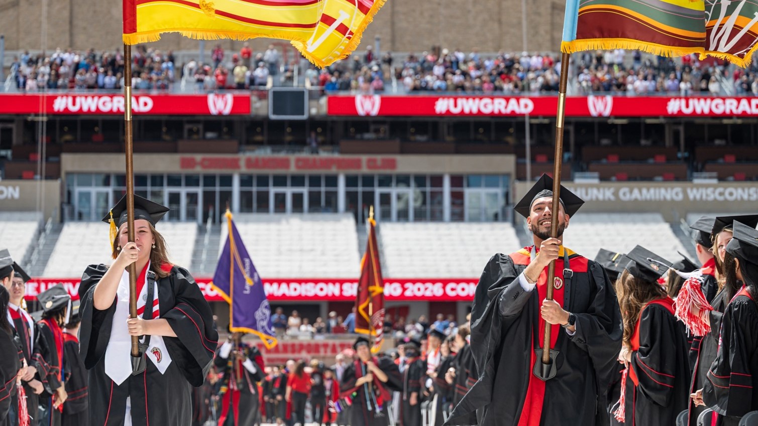 Two people carry flags in a large stadium. Both wear graduation robes.
