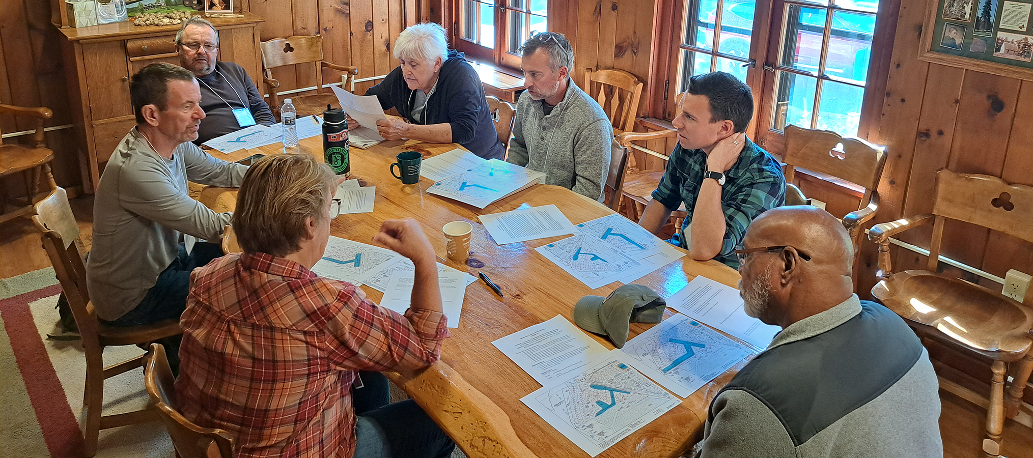 7 engaged adults sit at a wood table with lake maps spread out in front of them
