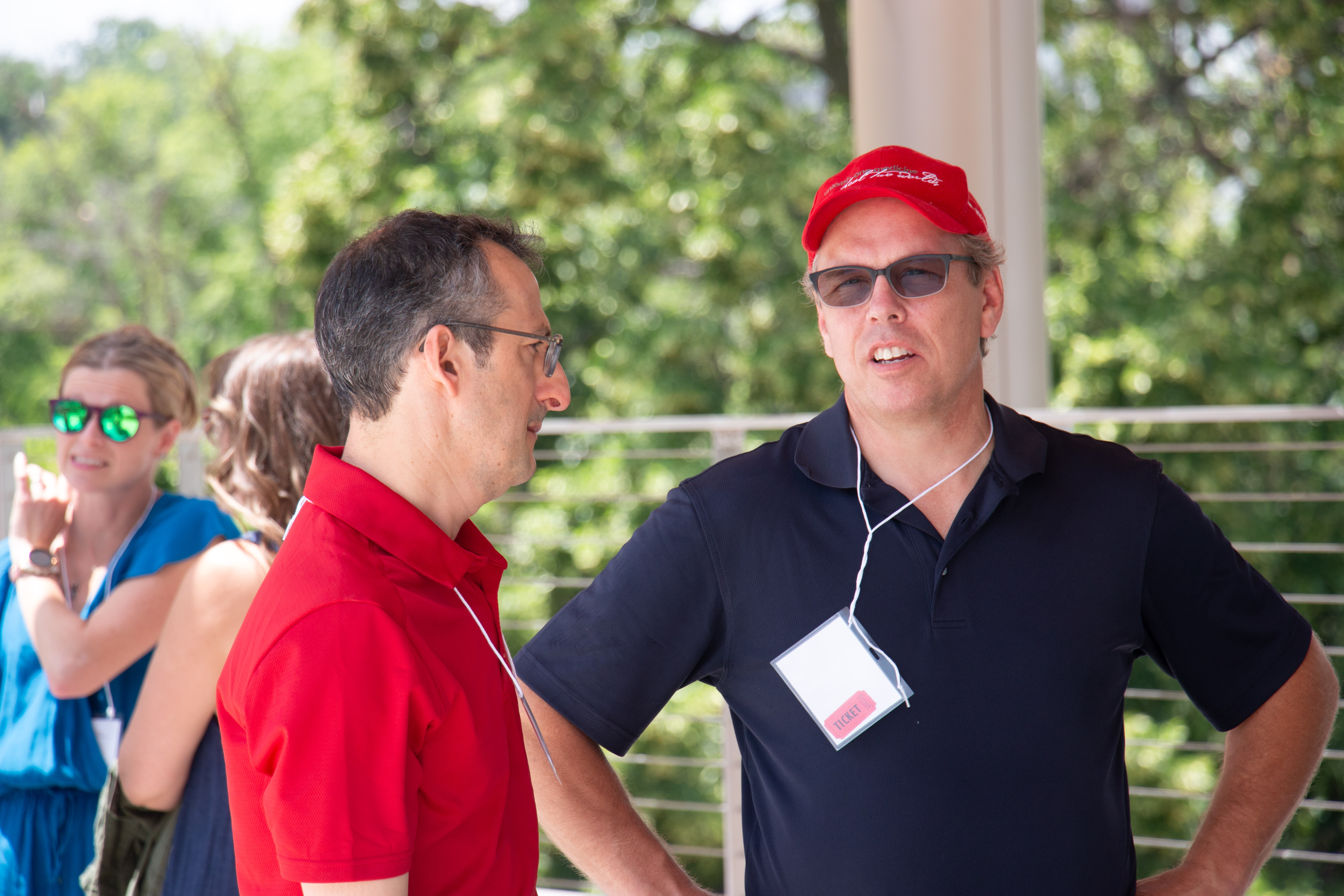 Two men conversing outdoors; one in red wearing glasses, the other in a hat and sunglasses. Green trees in background, giving a relaxed, sunny atmosphere.