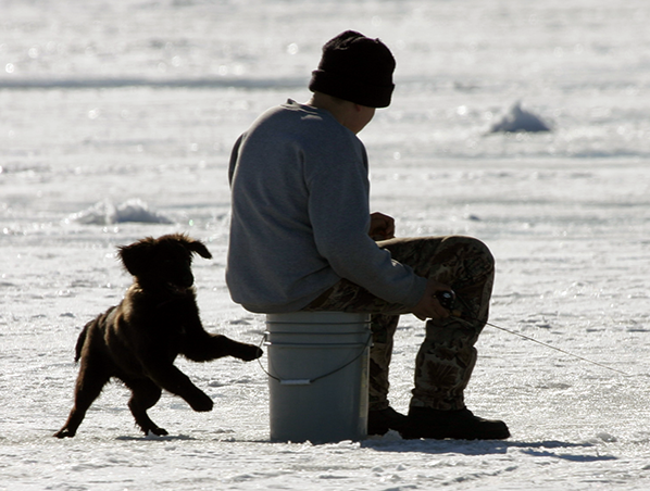 excited puppy next to a young boy sitting on a 5 gallon bucket while ice fishing