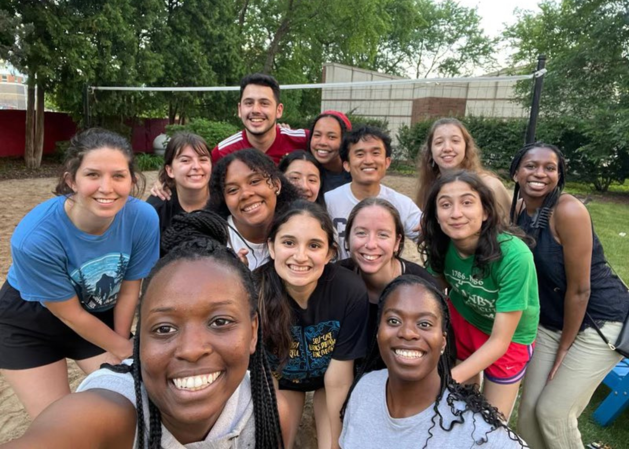 Fourteen young people taking a selfie on a sand volleyball court.