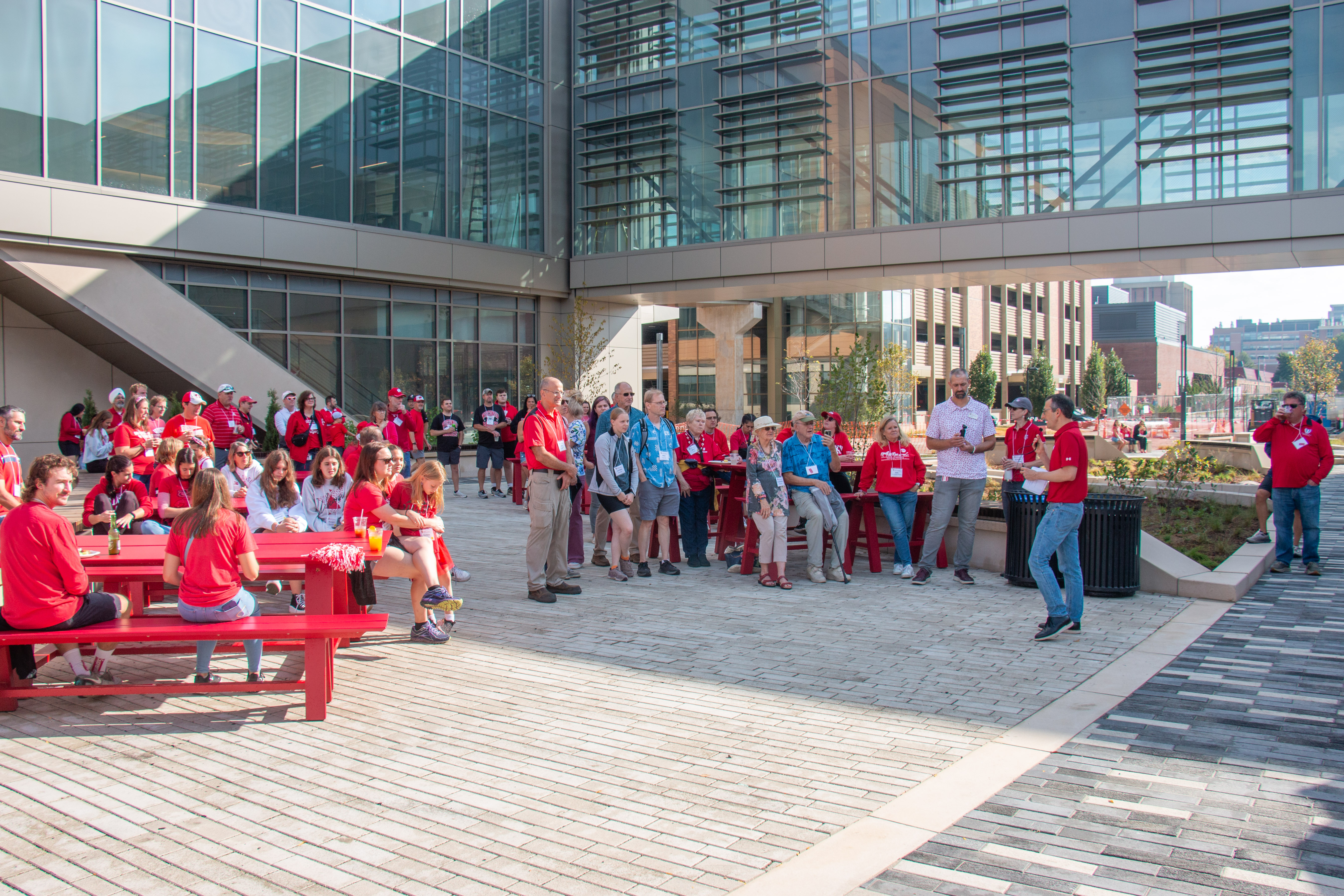 A group of people in red shirts gather outside a modern glass building. Some sit at red picnic tables, others stand, engaging and smiling. It's a sunny day.
