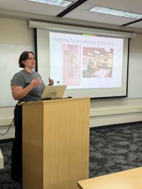 Lou Ginart stands at a lecture podium; in the background, slides show moments from her career