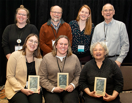 a group of people pose for a photo. Four people standing in the back and three people sit in front of them. The people who are sitting are holding blue tile awards. The background is a black curtain.