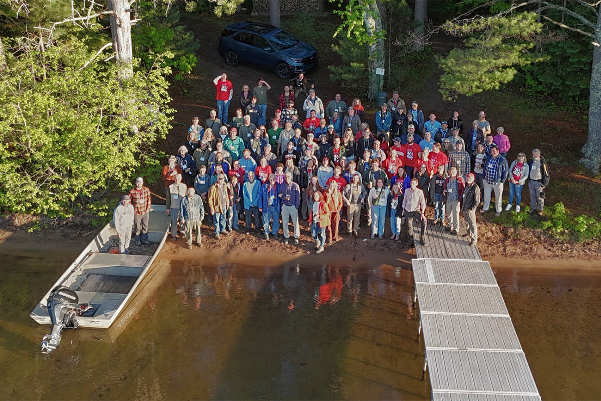 Drone Photo taken over lake facing shoreline of large group of people waving to drone.