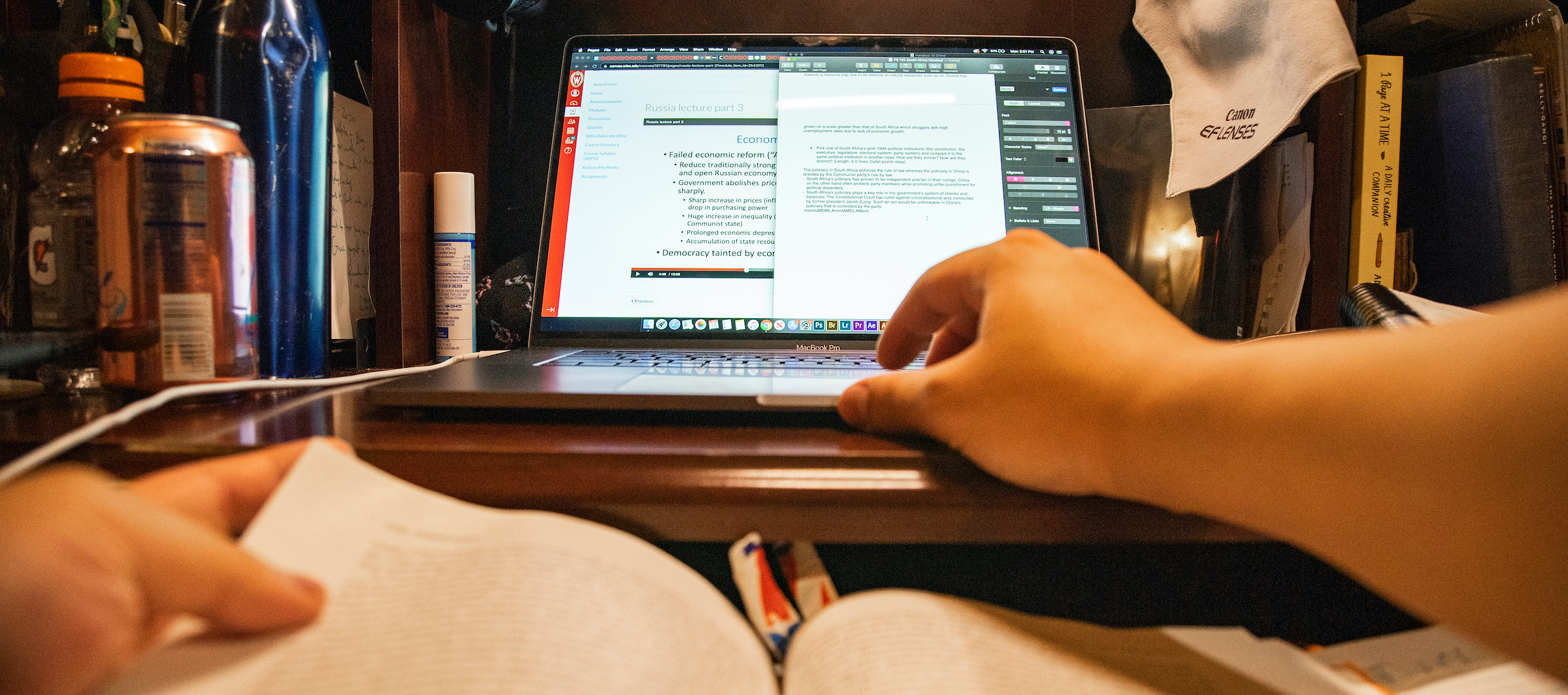 A laptop sits on a desk. Someone's right arm reaches toward the laptop, and their left had holds an open textbook in their lap.