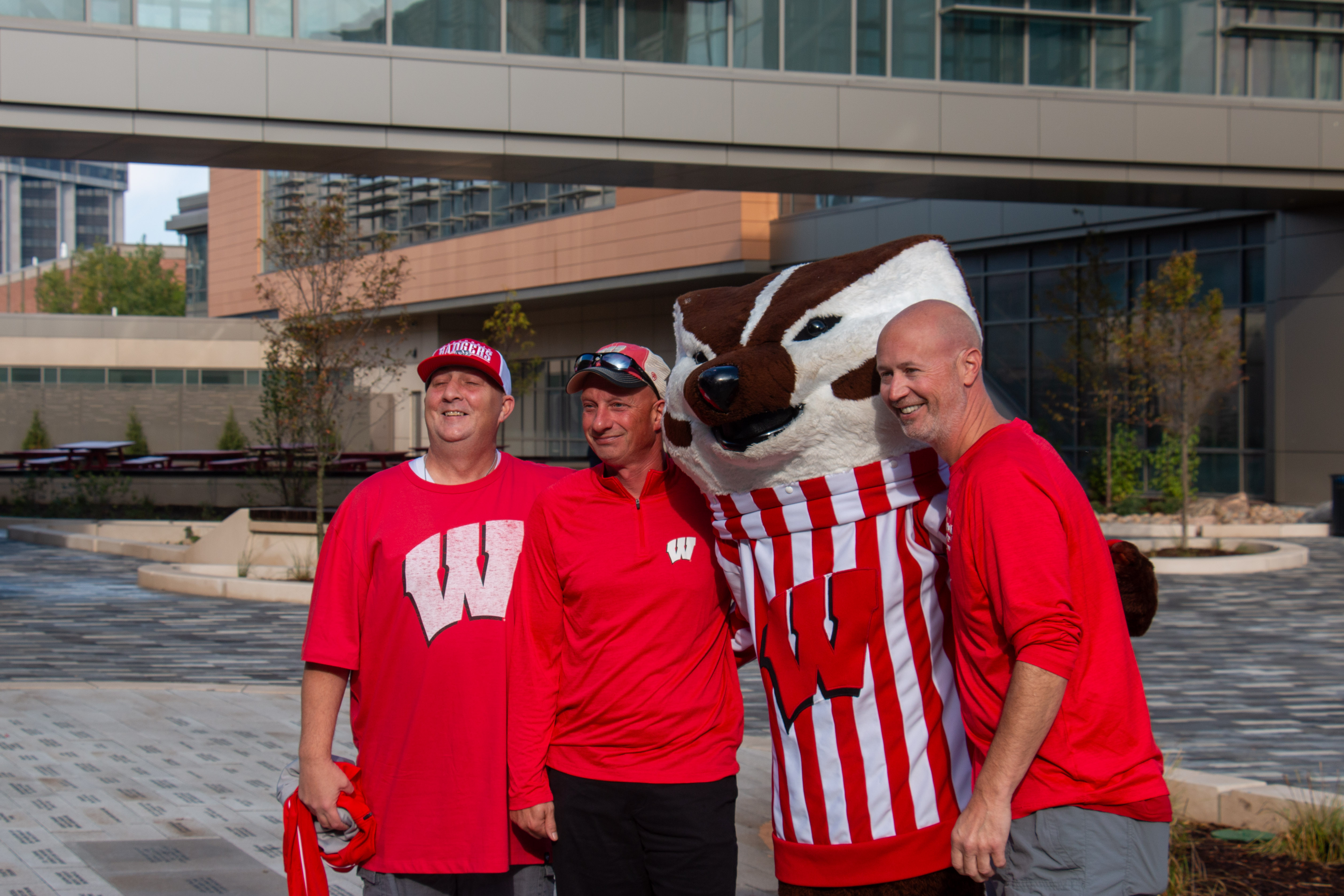 Three men in red Wisconsin Badgers gear and a large badger mascot pose happily outdoors in an urban setting, conveying school spirit and camaraderie.