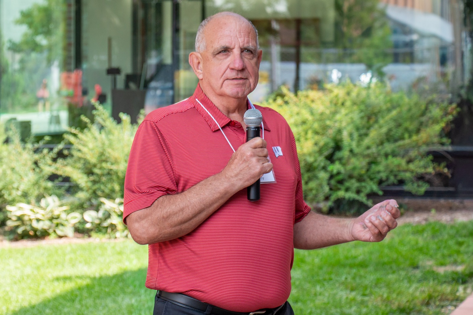 An older man in a red polo shirt speaks into a microphone outdoors. He stands on a lawn with greenery and a glass building in the background, projecting a thoughtful demeanor.