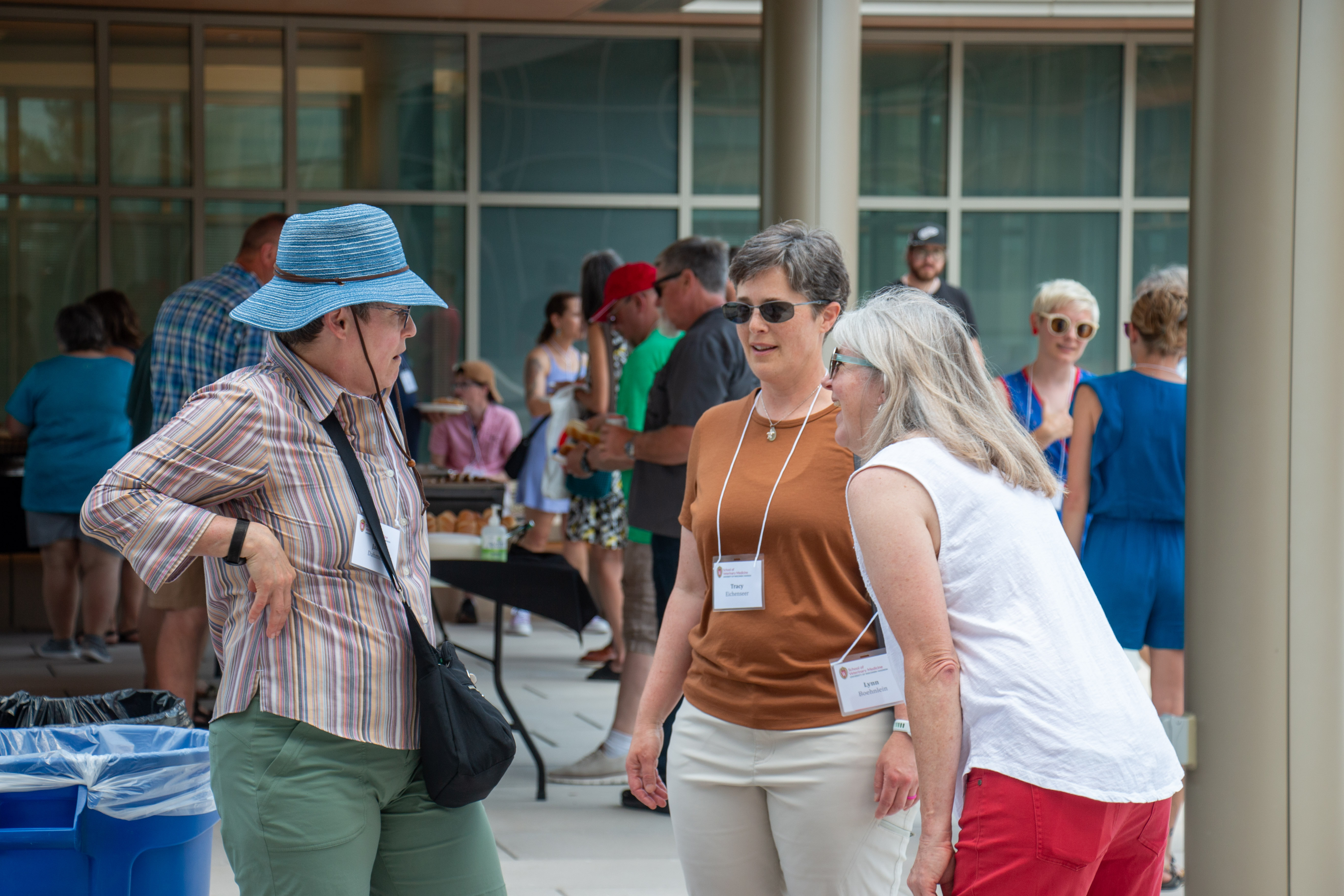 Three women engage in conversation outdoors, wearing casual summer attire. A blue hat and name tags are visible. People socialize in the background.
